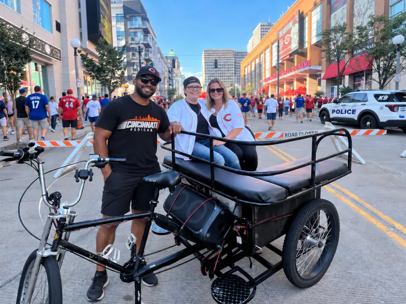 Pedicab driver with two Reds fans beside the cab on a busy game-day street